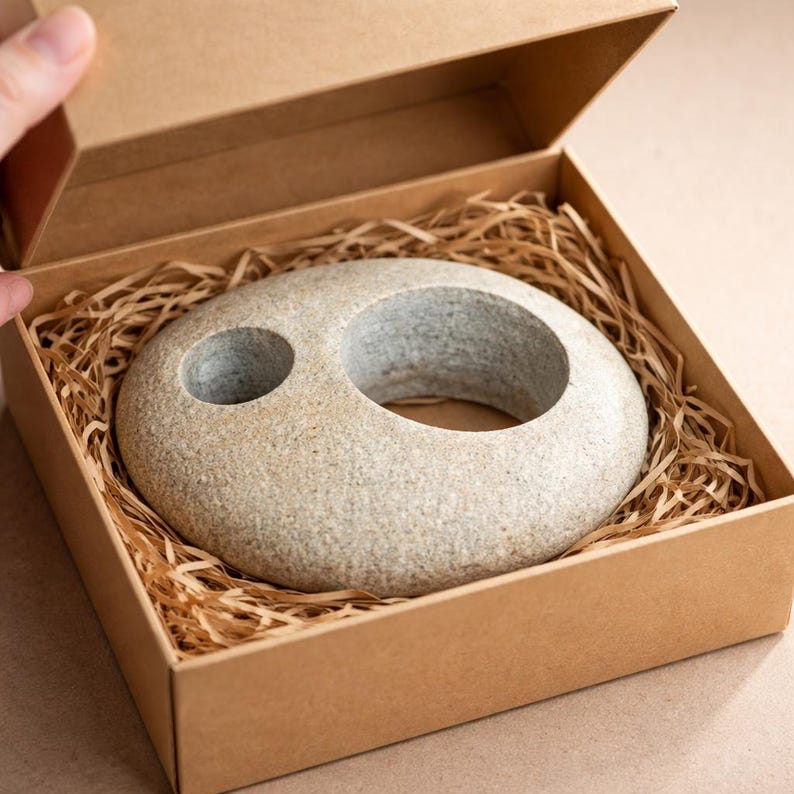 K&ouml;nnte beinhalten: Eine graue Steinskulptur mit einer gro&szlig;en kreisf&ouml;rmigen &Ouml;ffnung und einem kleineren Loch, pr&auml;sentiert in einer braunen Pappschachtel mit geschreddertem Papier. Die Skulptur hat eine glatte, abgerundete Form und eine nat&uuml;rliche, strukturierte Oberfl&auml;che.