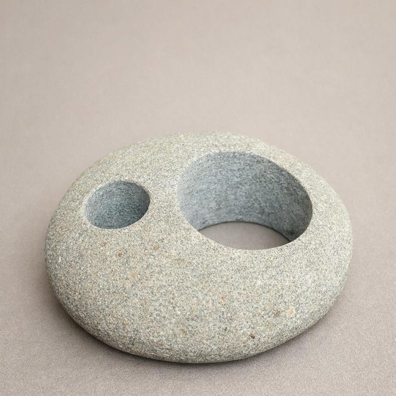 K&ouml;nnte beinhalten: Ein grauer Kerzenhalter aus Stein mit kreisf&ouml;rmigem Design. Er hat einen gro&szlig;en, offenen Kreis und ein kleineres Loch. Der Stein hat eine gesprenkelte Textur und ein nat&uuml;rliches Aussehen. Der Kerzenhalter ist ein dekorativer Artikel.