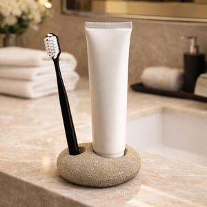 K&ouml;nnte beinhalten: Ein schwarzer Zahnb&uuml;rstenhalter mit wei&szlig;en Borsten und eine wei&szlig;e Tube Zahnpasta werden in einem beigefarbenen Steinhalter pr&auml;sentiert. Der Halter steht auf einer Marmorarbeitsplatte, was auf ein Badezimmer hindeutet. Die Zahnb&uuml;rste und Zahnpasta sind sauber und neu.