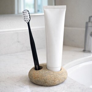 K&ouml;nnte beinhalten: Ein Halter aus Naturstein f&uuml;r Zahnb&uuml;rste und Zahnpasta. Er enth&auml;lt eine schwarze Zahnb&uuml;rste und eine wei&szlig;e Tube Zahnpasta. Der Halter steht auf einer wei&szlig;en Arbeitsplatte in einem Badezimmer.
