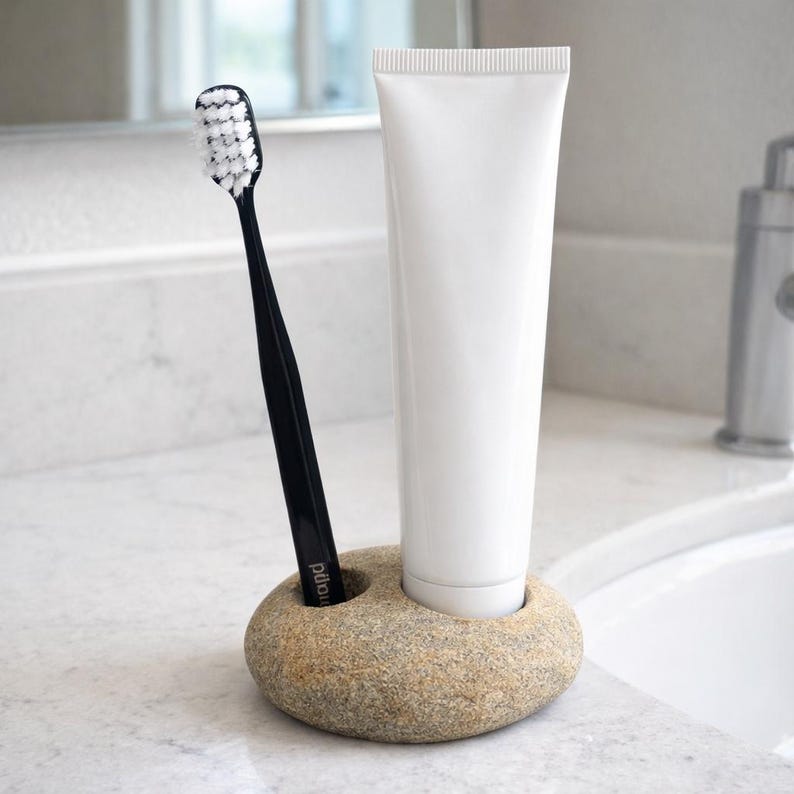 K&ouml;nnte beinhalten: Ein Halter aus Naturstein f&uuml;r Zahnb&uuml;rste und Zahnpasta. Er enth&auml;lt eine schwarze Zahnb&uuml;rste und eine wei&szlig;e Tube Zahnpasta. Der Halter steht auf einer wei&szlig;en Arbeitsplatte in einem Badezimmer.