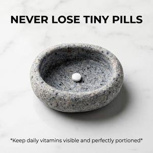 Puede incluir: Un pastillero de piedra gris de forma ovalada con una sola pastilla blanca en su interior. El texto "NEVER LOSE TINY PILLS" est&aacute; en la parte superior. El texto inferior dice "Keep daily vitamins visible and perfectly portioned."
