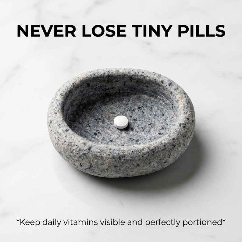 Puede incluir: Un pastillero de piedra gris de forma ovalada con una sola pastilla blanca en su interior. El texto "NEVER LOSE TINY PILLS" est&aacute; en la parte superior. El texto inferior dice "Keep daily vitamins visible and perfectly portioned."