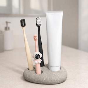 Può includere: Un portaspazzolini in pietra grigia con tre spazzolini e un tubetto di dentifricio. Gli spazzolini sono beige, neri e rosa, con setole nere e bianche. Lo spazzolino rosa ha un design da astronauta.