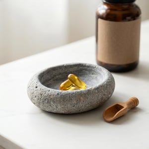 Puede incluir: Un cuenco de piedra gris contiene dos c&aacute;psulas amarillas. Una peque&ntilde;a cuchara de madera descansa cerca, con una botella de vidrio marr&oacute;n en el fondo. Los art&iacute;culos est&aacute;n dispuestos sobre una superficie blanca, sugiriendo un tema de salud y bienestar.
