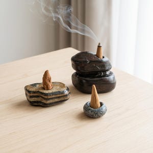 May include: Three stone incense holders of varying shapes and colors, each holding a tan cone-shaped incense. One holder is dark brown and stacked, one is striped with black and tan, and one is small, round, and grey speckled with smoke rising.