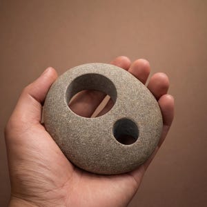 K&ouml;nnte beinhalten: Eine graue Steinskulptur mit zwei kreisf&ouml;rmigen L&ouml;chern, einem gro&szlig;en und einem kleinen. Der glatte, oval geformte Stein wird in einer Hand vor einem braunen Hintergrund gehalten. Die Skulptur hat eine nat&uuml;rliche, organische &Auml;sthetik.