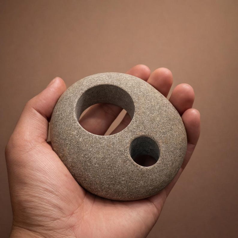 K&ouml;nnte beinhalten: Eine graue Steinskulptur mit zwei kreisf&ouml;rmigen L&ouml;chern, einem gro&szlig;en und einem kleinen. Der glatte, oval geformte Stein wird in einer Hand vor einem braunen Hintergrund gehalten. Die Skulptur hat eine nat&uuml;rliche, organische &Auml;sthetik.