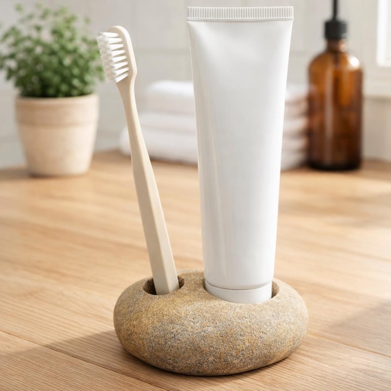 K&ouml;nnte beinhalten: Ein beiger Zahnb&uuml;rste und eine wei&szlig;e Zahnpasta-Tube in einem Steinhalter. Der Halter hat zwei L&ouml;cher, eines f&uuml;r die Zahnb&uuml;rste und eines f&uuml;r die Zahnpasta. Im Hintergrund sind eine Pflanze und eine braune Flasche zu sehen.