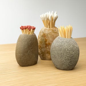 Può includere: Tre contenitori decorativi in pietra che contengono fiammiferi, cotton fioc e stuzzicadenti. I contenitori sono in tonalità marroni e grigie, con un aspetto strutturato e organico. I fiammiferi hanno punte rosse.