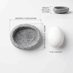 Puede incluir: Un cuenco de piedra gris de forma ovalada, de 5-8 cm de largo, 4-6 cm de ancho y 2-4 cm de alto. Un huevo blanco se encuentra junto al cuenco para comparaci&oacute;n de escala.