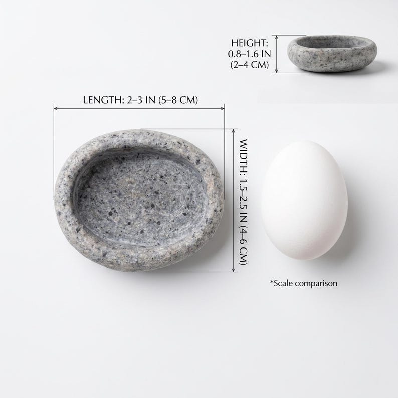 Puede incluir: Un cuenco de piedra gris de forma ovalada, de 5-8 cm de largo, 4-6 cm de ancho y 2-4 cm de alto. Un huevo blanco se encuentra junto al cuenco para comparaci&oacute;n de escala.