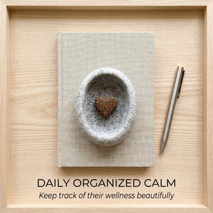 Puede incluir: Una composici&oacute;n de tonos neutros con un libro, un cuenco de piedra con un objeto en forma de coraz&oacute;n y un bol&iacute;grafo. El texto "DAILY ORGANIZED CALM" y "Keep track of their wellness beautifully" se muestra en la parte inferior. Los objetos est&aacute;n dispuestos en una bandeja de madera.