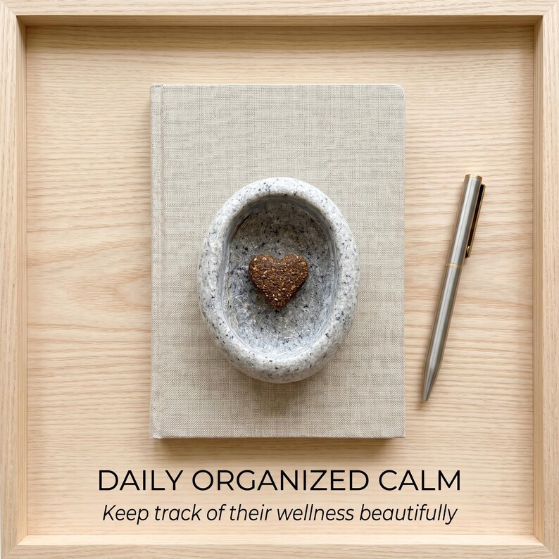 Puede incluir: Una composici&oacute;n de tonos neutros con un libro, un cuenco de piedra con un objeto en forma de coraz&oacute;n y un bol&iacute;grafo. El texto "DAILY ORGANIZED CALM" y "Keep track of their wellness beautifully" se muestra en la parte inferior. Los objetos est&aacute;n dispuestos en una bandeja de madera.