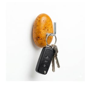May include: A unique key holder featuring a potato-shaped design with a metal hook. A set of keys, including a black car key fob, hangs from the hook. The potato has a textured, brown surface, set against a white background.