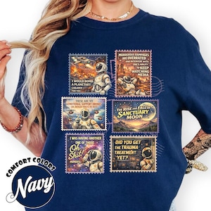 May include: Navy blue t-shirt with several postage stamp-style graphics featuring astronauts and space-themed text. The stamps include phrases such as "I would bomb a planetary colony for you" and "Did you get the trauma treatment yet?"