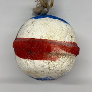 May include: A spherical white foam fishing float featuring a prominent horizontal red band around the center and blue painted sections at the top and bottom. A frayed, light-colored rope loop is attached to the top of the textured, weathered surface.