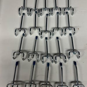 May include: A collection of twenty silver-colored metal pegboard hooks arranged in four rows of five. Each hook features a double-pronged base for mounting and a single long arm extending outward, topped with a small blue plastic protective cap.