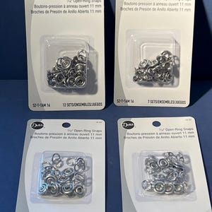 May include: Four white retail blister packs of Dritz 7/16 inch open-ring snaps. Each package contains silver-colored metal fasteners visible through a clear plastic bubble. The text on the cards reads Dritz, 7/16 inch Open-Ring Snaps, and SZ-T-TAM 16.