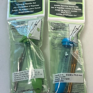 May include: Two Clover brand darning needle sets in clear plastic packaging. One set has a green cap, the other blue. Each package contains needles and a plastic tube. Text on the packaging includes "Darning Needle Set".