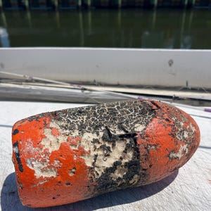 May include: An orange and black buoy with a weathered appearance. The buoy has a cylindrical shape with orange paint on the ends and black, cracked paint in the middle. The buoy is likely used for marking locations in the water.