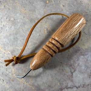 May include: A leathercraft stitching groover tool featuring a polished light brown wooden handle with a ribbed grip section, a short metal hook tip, and a thin tan leather cord tied in a loop attached to the end of the handle.