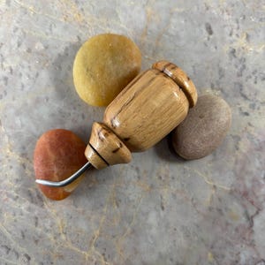 May include: A seam ripper featuring a polished, light-brown wooden handle with a distinct grain pattern and a curved metal blade. The tool is positioned among three smooth, rounded stones in shades of yellow, orange, and grey.