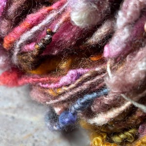 May include: A skein of art yarn featuring a textured blend of fibers in shades of magenta, deep pink, mustard yellow, brown, and soft blue. The yarn displays varying thicknesses, including fuzzy, coiled, and smooth sections throughout the strand.