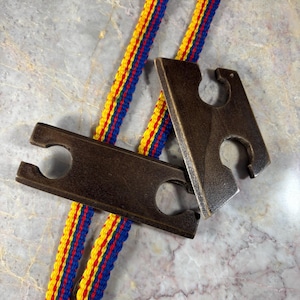 May include: Two dark brown wooden rectangular pieces with rounded cutouts on the sides, positioned over a long, thin woven strap featuring a repeating pattern of vertical yellow, red, and blue stripes running along its entire length.