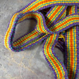 May include: A woven fabric strap featuring a repeating pattern of vertical stripes in purple, orange, yellow, and teal. The textile band is flexible and folded, displaying a consistent weave structure with visible thread textures throughout the length.