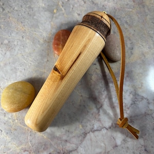 May include: A cylindrical wooden handle with a light tan grain and a dark brown textured top section. A thin leather cord is attached to the top, featuring a knot at the end. The object is positioned near three smooth, rounded stones.
