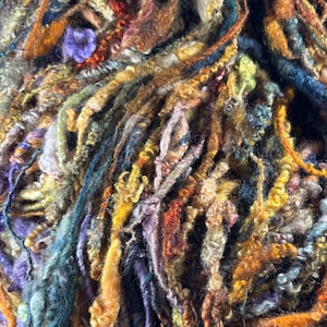 May include: A multi-textured art yarn featuring a complex blend of fibers in shades of burnt orange, deep teal, mustard yellow, lavender, and earthy brown. The yarn displays varied thicknesses, including thin strands, thick coils, and fuzzy loops.