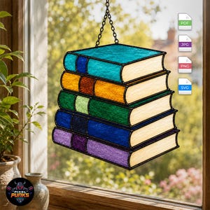 Puede incluir: Un atrapasoles de vidrieras que muestra una pila de cinco libros en colores turquesa, naranja, verde, azul y morado. Cada libro tiene un lomo texturizado y páginas de color crema. La pieza cuelga de una cadena de metal negro ante una ventana.