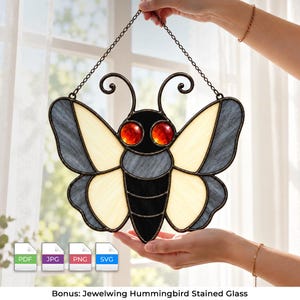 May include: A stained glass suncatcher shaped like a moth with a black body, cream-colored wings, and two glowing red eyes. The piece is framed in dark metal and hangs from a thin chain. Below the image are four file icons labeled PDF, JPG, PNG, and SVG.