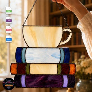 Puede incluir: Un atrapasoles de vidrieras que muestra una taza de té color crema sobre una pila de tres libros. Los libros tienen lomos en azul oscuro, rojo intenso y morado oscuro con portadas claras. La pieza está suspendida por una cadena de metal.