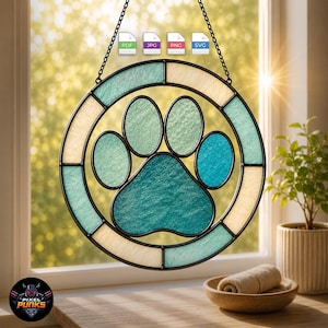 May include: A circular stained glass suncatcher featuring a central paw print design in shades of teal and blue, surrounded by a border of alternating cream and teal glass segments. The piece is suspended by a black metal chain for hanging.