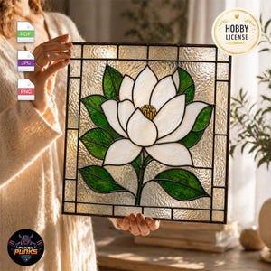 Op de afbeelding: Een vierkant glas-in-loodpaneel met een centrale witte magnoliabloem met gele meeldraden en donkergroene bladeren. Het ontwerp is omlijst door een donker metalen raster tegen een getextureerde, doorschijnende achtergrond.