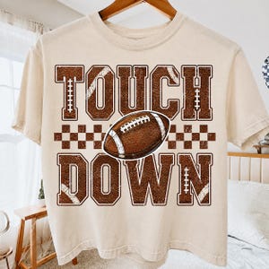Könnte beinhalten: Ein beiges Kurzarm-T-Shirt mit einem braunen Grafikdruck, der den Text TOUCH DOWN in einer abgenutzten College-Schrift zeigt. Ein brauner Football mit weißen Schnürsenkeln ist zwischen den Wörtern zentriert, darunter befindet sich ein Karomuster.
