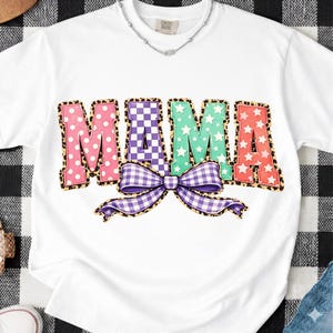 Peut inclure: T-shirt blanc avec le mot "MAMA" en lettres colorées et à motifs. Les lettres sont bordées d'un imprimé léopard. Un nœud vichy violet et blanc est en dessous. Un collier argenté est visible au niveau du col.