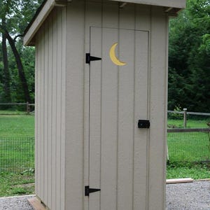 Può includere: Una piccola casetta di legno beige con tetto spiovente. La porta presenta un ritaglio a forma di luna crescente gialla. Cerniere nere e un chiavistello fissano la porta. La casetta è su una base di ghiaia, circondata dal verde.
