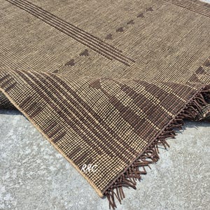 May include: A woven brown rug with a geometric pattern. The rug features a tan background with dark brown stripes and triangle shapes. The edge of the rug is folded over, revealing a fringe.