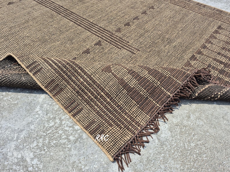 May include: A woven brown rug with a geometric pattern. The rug features a tan background with dark brown stripes and triangle shapes. The edge of the rug is folded over, revealing a fringe.