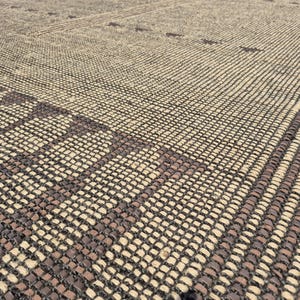 May include: Close-up of a woven rug with a geometric pattern. The rug features a combination of beige and dark brown tones, creating a textured surface. The design includes rectangular and striped elements.