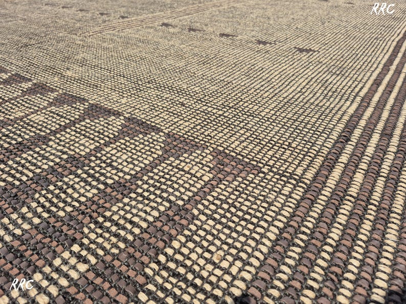 May include: Close-up of a woven rug with a geometric pattern. The rug features a combination of beige and dark brown tones, creating a textured surface. The design includes rectangular and striped elements.
