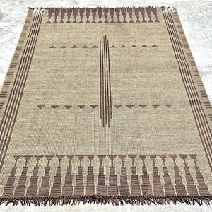 May include: A rectangular beige rug with a geometric pattern. The rug features a central vertical stripe and horizontal rows of triangles and stylized shapes in brown. The edges have a striped border and fringe.