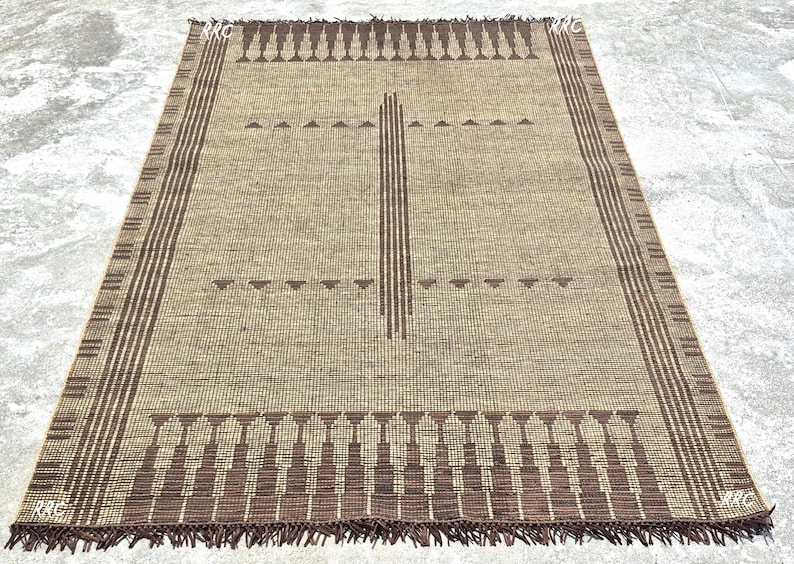 May include: A rectangular beige rug with a geometric pattern. The rug features a central vertical stripe and horizontal rows of triangles and stylized shapes in brown. The edges have a striped border and fringe.
