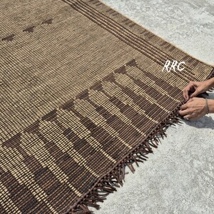 May include: A woven brown and tan rug with a geometric pattern and fringe detail. The rug features a grid-like design with darker brown accents and a decorative border. The rug is made of natural fibers and is suitable for indoor use.