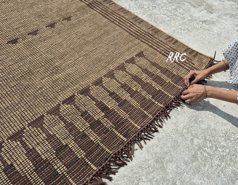 May include: A woven brown and tan rug with a geometric pattern and fringe detail. The rug features a grid-like design with darker brown accents and a decorative border. The rug is made of natural fibers and is suitable for indoor use.