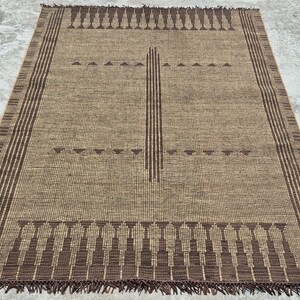 May include: A rectangular, woven rug in shades of brown and tan. The rug features a central vertical design and borders with geometric patterns. The edges have a fringe detail. The letters "RRC" are visible in the bottom left corner.