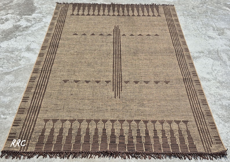 May include: A rectangular, woven rug in shades of brown and tan. The rug features a central vertical design and borders with geometric patterns. The edges have a fringe detail. The letters "RRC" are visible in the bottom left corner.